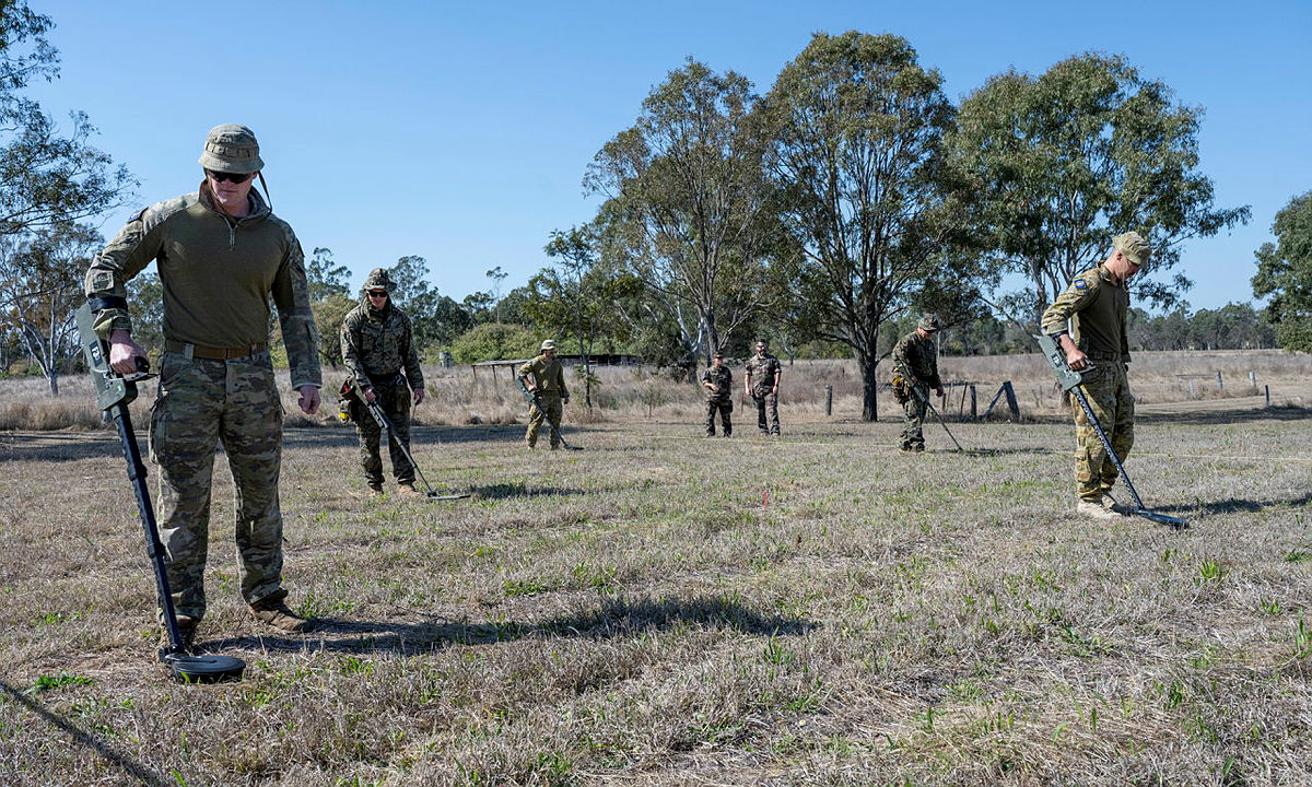 Seven uniformed personnel from the Australian Defence Force use metal detectors to search for potentially dangerous war remnants in a field in Nauru. They are deployed as members of the ordnance mission Operation Render Safe.