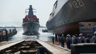 Turkey's new offshore patrol vessels, AKHİSAR ( P-1220) and KOÇHİSAR (P-1221) are seen on a dock, with P-1220 already on the water and P-1221 on the ready for launch. Dock workers can be seen on the ground, by the side of P-1221. Both ships are adorned with wreaths and flags of Turkey.
