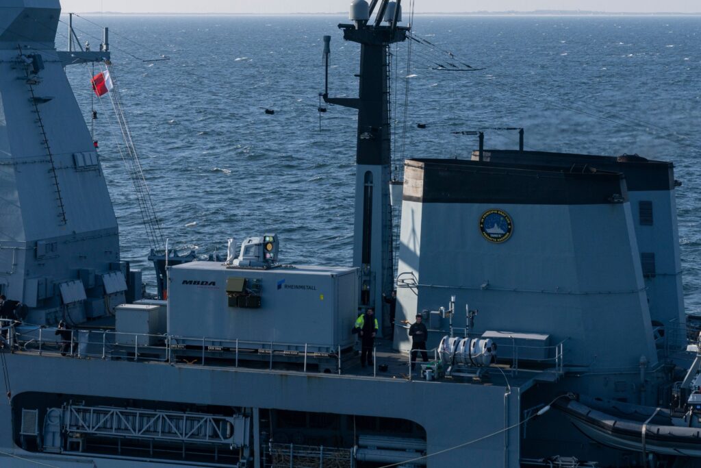 Part of the upper deck of Germany's Sachsen F-124 ship is seen on the photo. the laser weapon demonstrator, which looks like a giant rectangular box, is seen on the center left. The initials "MBDA" is written on its left side, and the name "Rheinmetall" is on the right. The sea, stretching over the horizon, serves as the background of the picture.