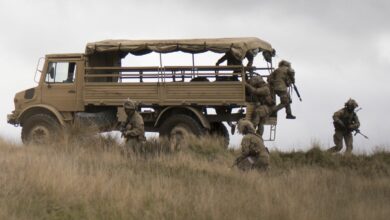 A stationary Mercedes Benz Unimog is seen in a grassland. Around its back, three camouflaged soldiers surround it and three more are jumping out of it.