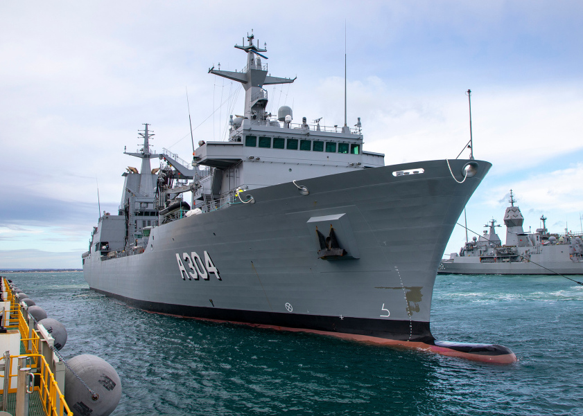 HMAS Stalwart, a Royal Australian Navy ship, is seen docked in a pier. It faces to the right, and "A304" is seen painted on its right side. On the middle right side of the background, another ship can be seen docked in the pier.