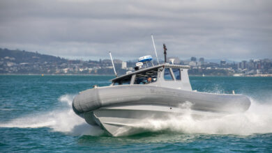 A Royal New Zealand Navy Littoral Manoeuvre Craft boat is seen gliding across the waters. A mountainside is seen in the back.