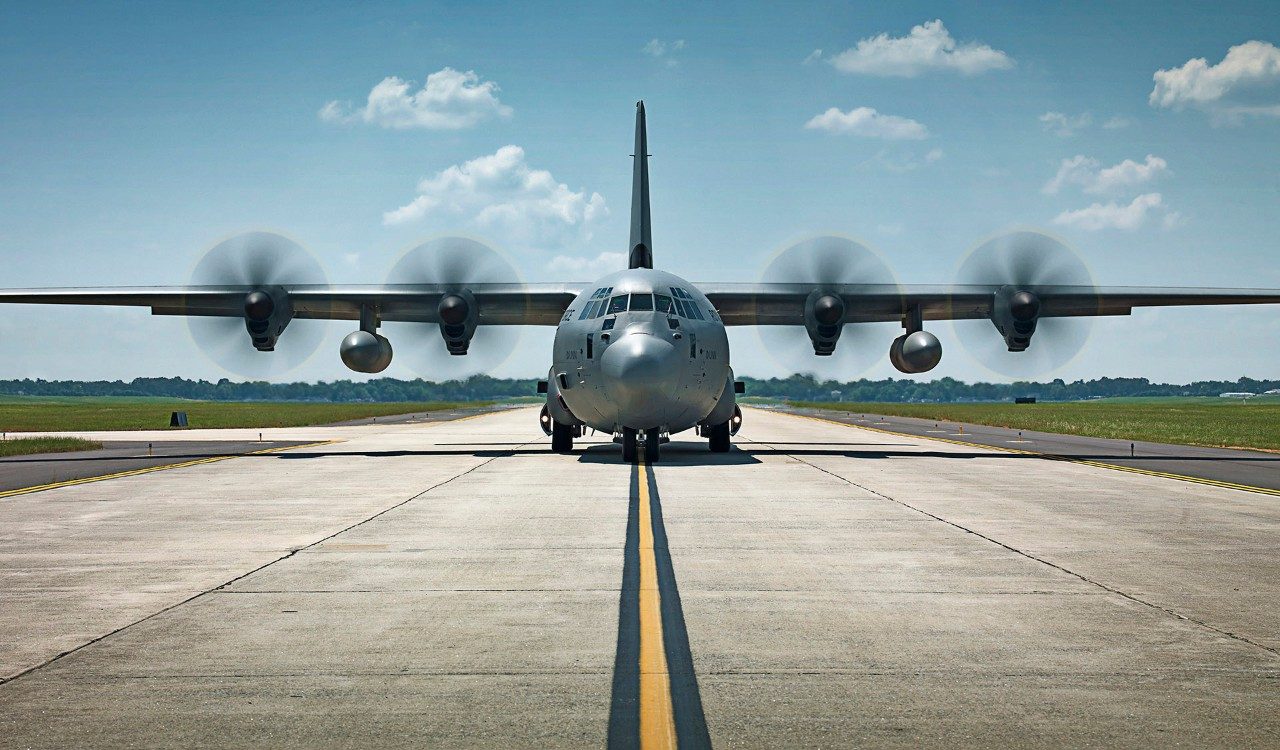 A Lockheed Martin C-130J-30 Super Hercules plane is seen front and center on the image. Its four wing propellers are spinning as it appears to be stationary on an airplane runway.