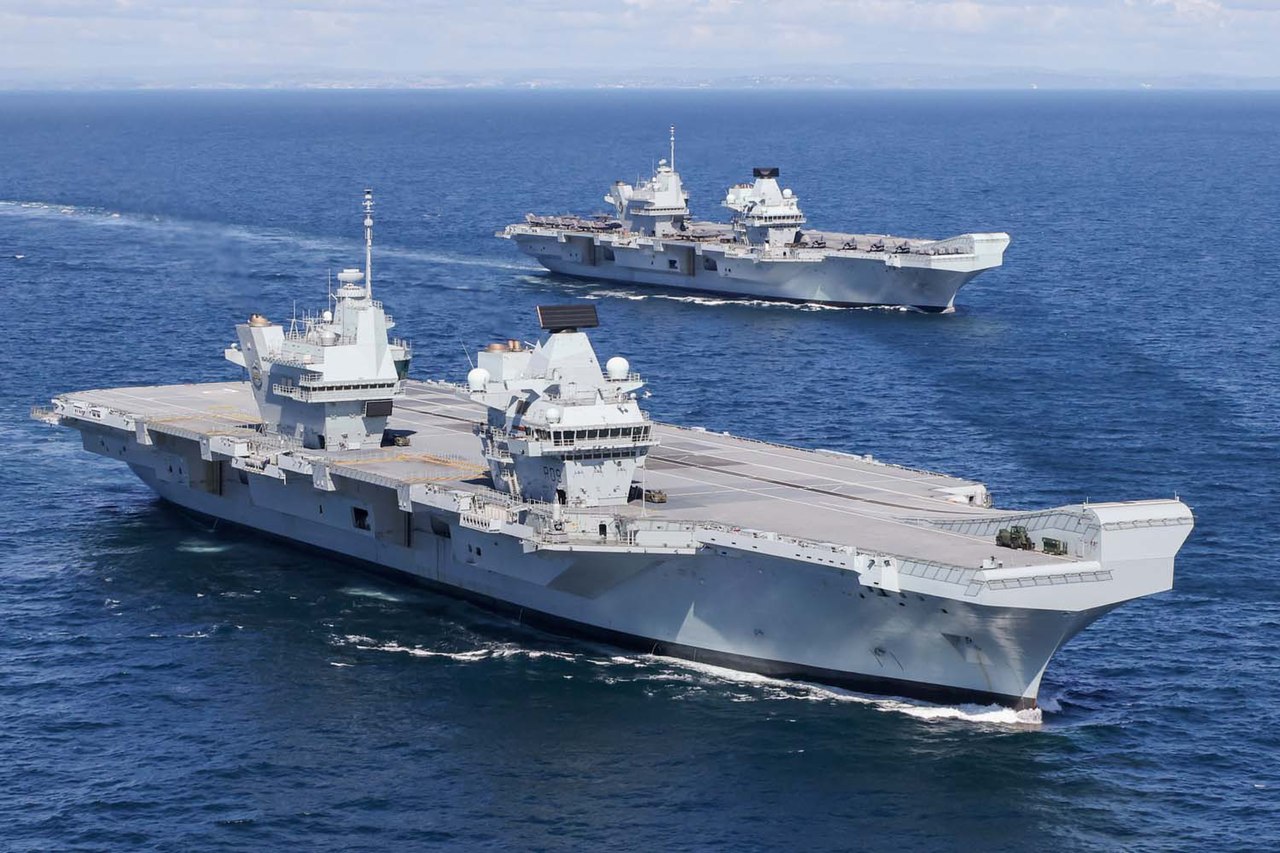 HMS Prince of Wales and HMS Queen Elizabeth pictured at sea. The water is a deep blue, and the cloudy sky serves as the background.