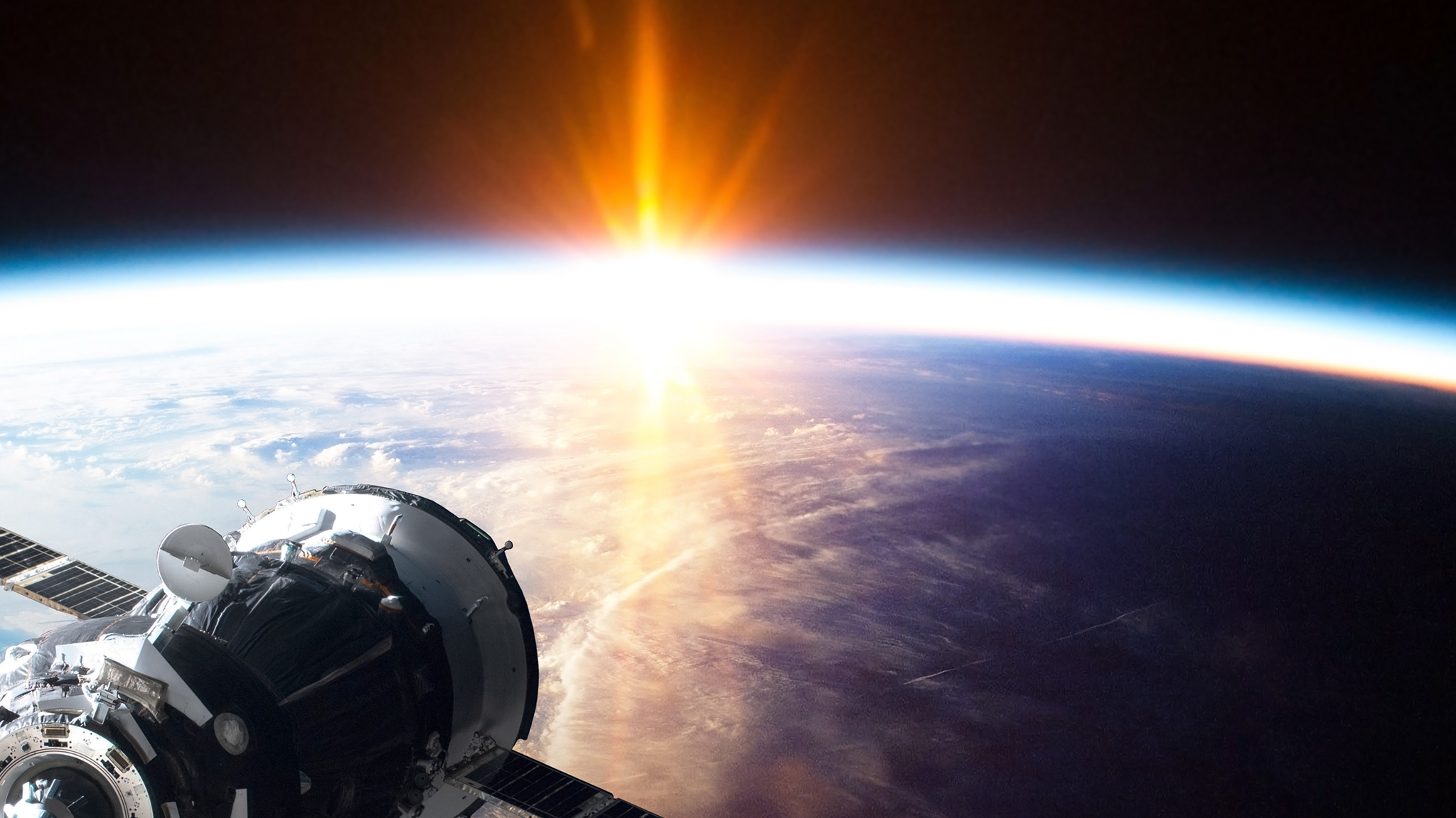 A satellite is seen in space, on the lower right corner of the image, The earth is seen on the background, and the sun is rising on its horizon.