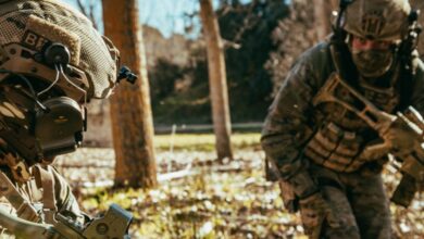 Two infantry soldiers in camouflage gear talk in a field with trees.