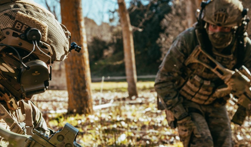 Two infantry soldiers in camouflage gear talk in a field with trees.