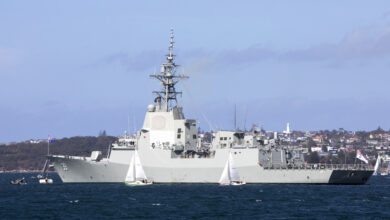 HMAS Hobart in Sydney Harbour prior to departing for her Mariner Skills Evaluation period. It has a white-grey hull with the number '39' painted on its left side in white. A coastal town serves as the background. The water is a deep blue.