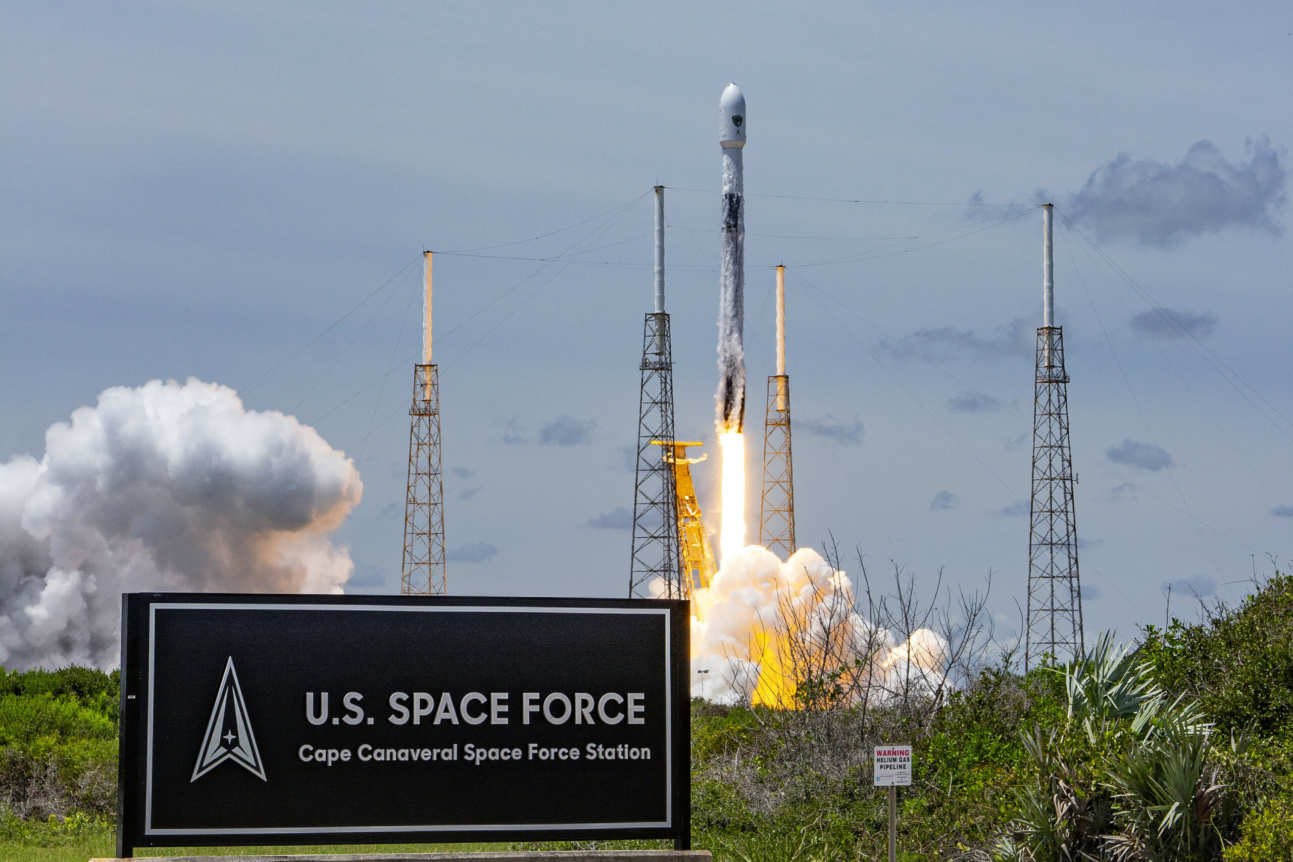 A rocket launches from at Cape Canaveral Space Force Station, Florida. On the left of the foreground, a sign that says "U.S. SPAC FORCE Cape Canaveral Space Force Station" is seen. The Space Force Logo is on the left of the sign.