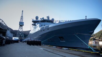 KV Bjørnøya (W311) is seen docked at Vard Langsten shipyard. Its grey hull has "W 311" painted on its right side in black. A stair that leads up to the deck of the ship juts out a little farther back its right side, with "BJØRNØYA" written on a big white fabric covering the stairs' side railings. Just a little closer to the foreground from it, lines of uniformed naval personnel are standing at attention.