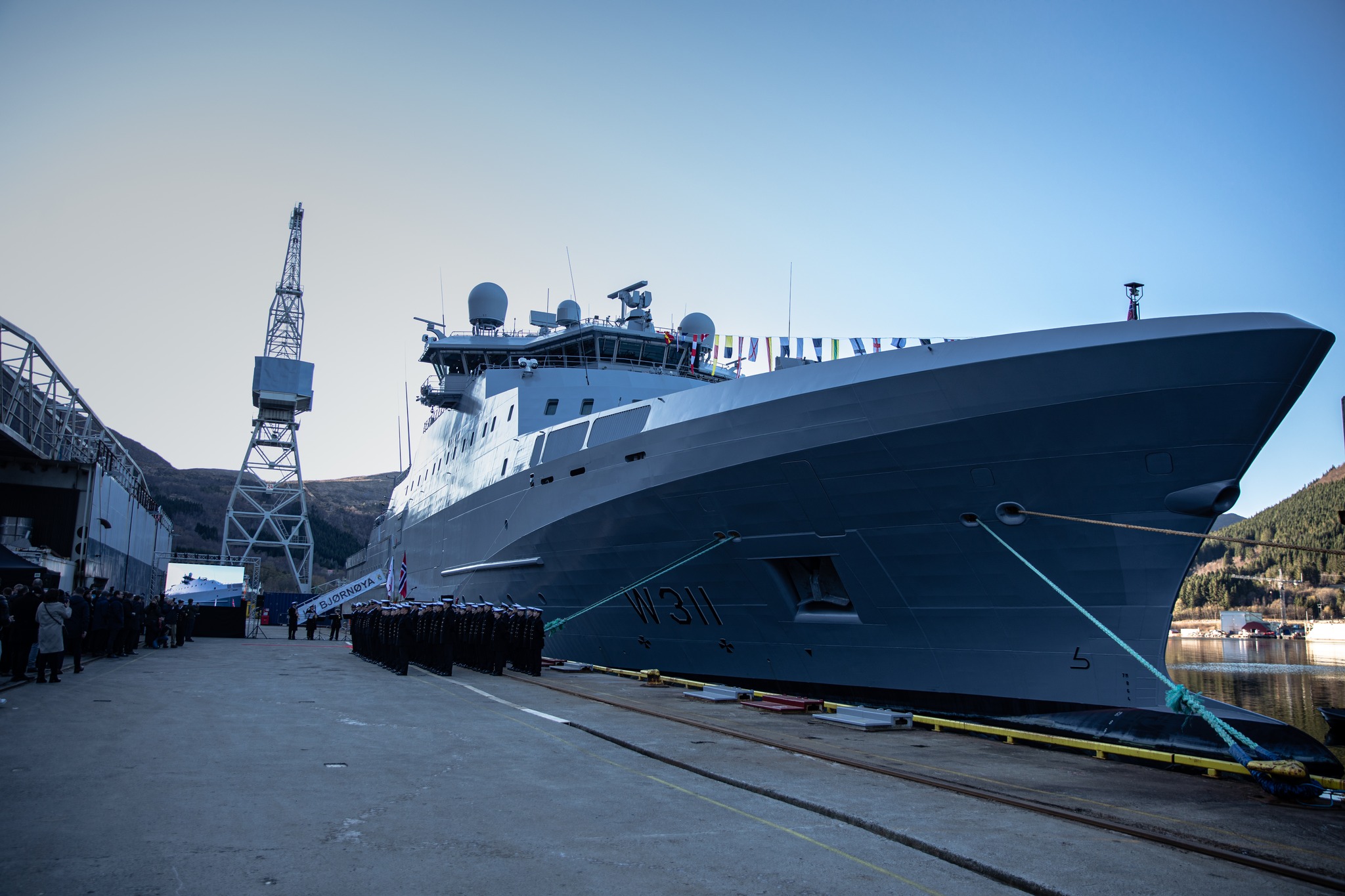 KV Bjørnøya (W311) is seen docked at Vard Langsten shipyard. Its grey hull has "W 311" painted on its right side in black. A stair that leads up to the deck of the ship juts out a little farther back its right side, with "BJØRNØYA" written on a big white fabric covering the stairs' side railings. Just a little closer to the foreground from it, lines of uniformed naval personnel are standing at attention.