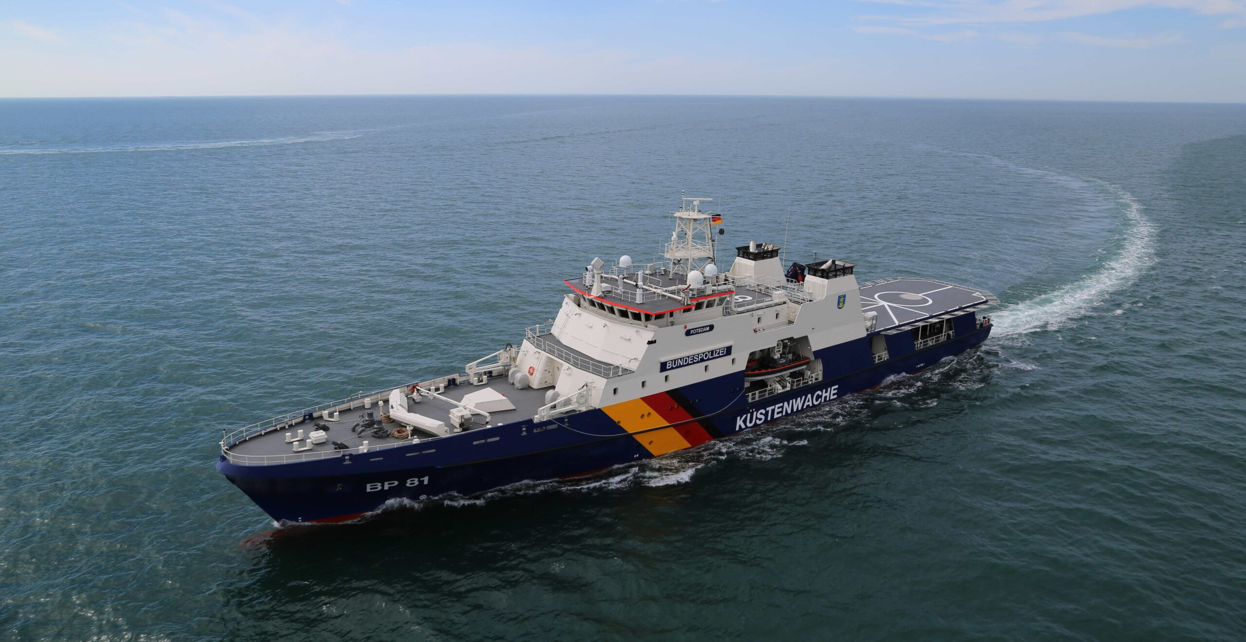 The German Federal Police's offshore patrol vehicle, Potsdam (BP81), sails in the deep blue-green sea. A relatively fair but hazy sky serves as the far background. The middle of its side has the word "KÜSTENWACHE" painted in white; "BP81" is seen painted right by the front as well.