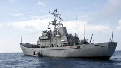 The Bangladesh Navy Ship Sangu (P-713) sails on the water. The ship is painted grey and it has "P713" painted on its front and side in black. A clear blue sky acts as the background, with a few white clouds along the middle. The foreground is the sea, with a deep blue color.