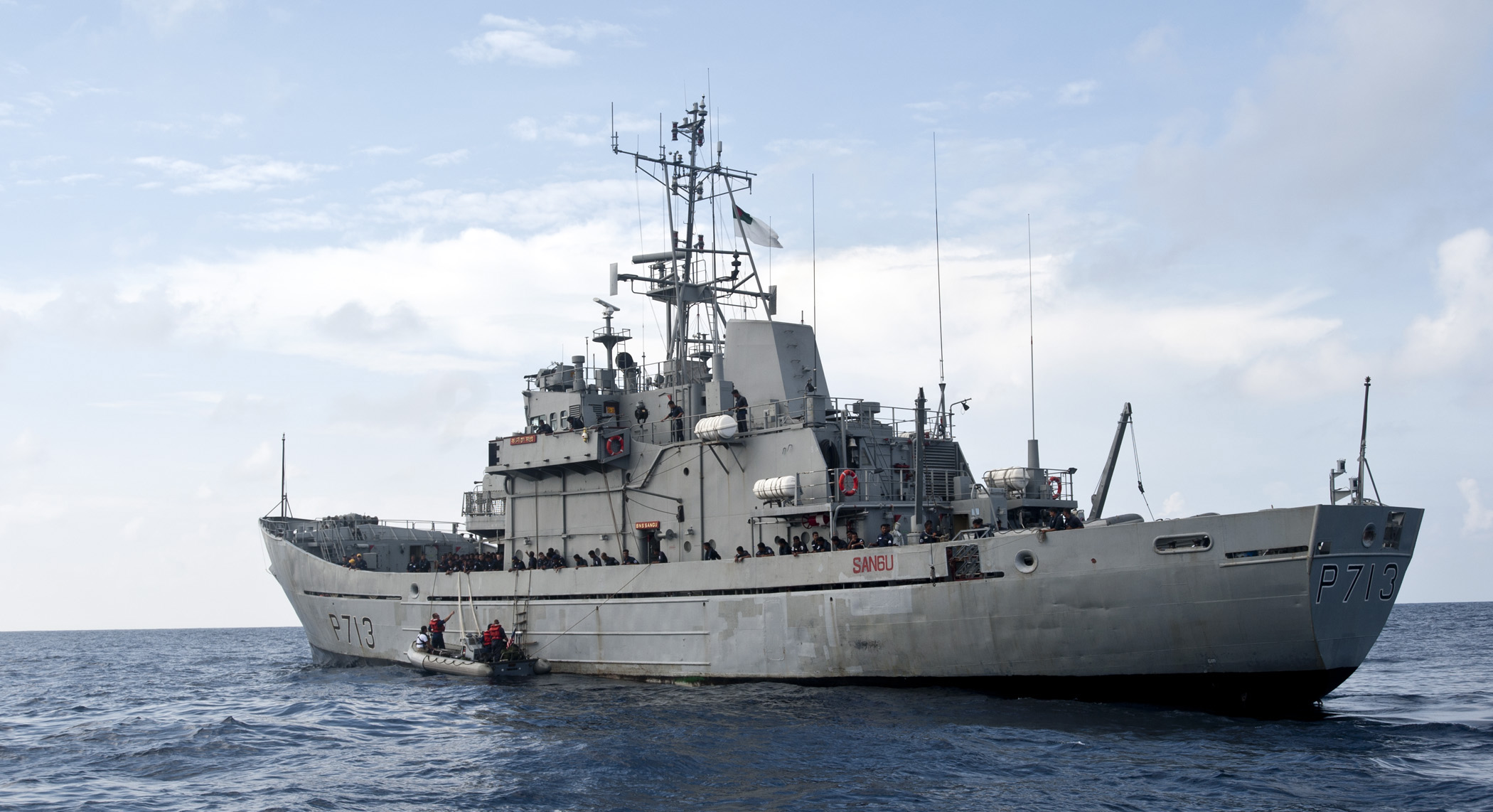 The Bangladesh Navy Ship Sangu (P-713) sails on the water. The ship is painted grey and it has "P713" painted on its front and side in black. A clear blue sky acts as the background, with a few white clouds along the middle. The foreground is the sea, with a deep blue color.