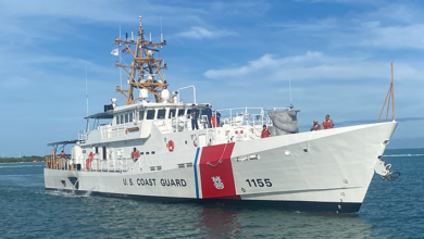 The US Coast Guard's Sentinel-class cutter, USCGC Melvin Bell (WPC-1155), is seen sailing the water. The ship is painted white, with the words "U.S. COAST GUARD" painted on the side of the hull in black. Close to the front, "1155" is also painted. Separating the two are a thin stripe of blue and a thick stripe of red, where the coast guard's coat of arms is located. Nine crew members are seen on deck. A fair, sparsely-cloudy blue sky serves as the image's background. The water is a deep blue-green color.