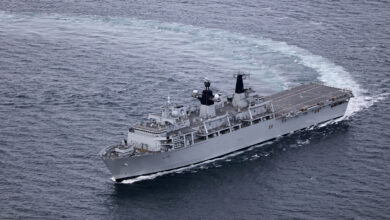 HMS Albion, an amphibious warfare ship, is seen sailing blue-grey waters. The photo appears to have been taken from above the waters, possible from an aircraft. The ship is painted grey and has "L14" painted on its side in black.