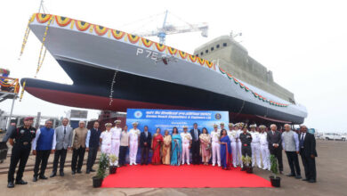 ASW-SWC Amini (P75), an Arnala-class anti-submarine corvette, is seen docked in the background. The photo was taken during its launching ceremony, and a group of 28 people are lined up in the foreground. They appear to be officials from the Indian Navy and the shipyard the corvette was built in. A red carpet is laid out in the middle and a banner for the event is displayed behind them.