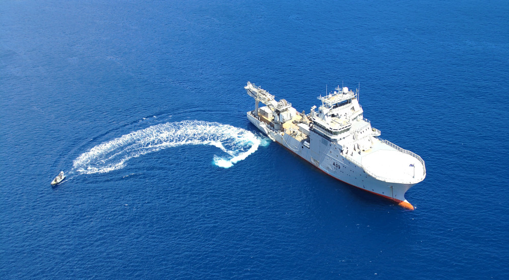 HMNZS Manawanui (A09) is seen sailing in the water as a smaller seacraft sails from it to the left. "A09" is painted in black on the right side of its grey hull.