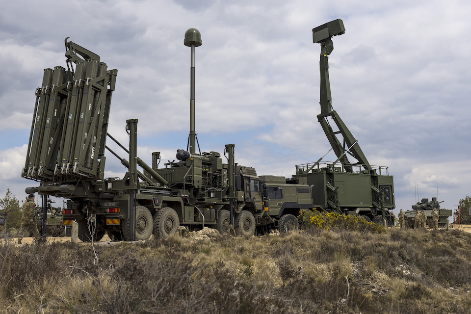 Sky Sabre, a state-of-the-art air defence system, is seen stationed in a grassy plain. Two trucks face each other, carrying various radar/sensor-looking equipment on their backs. On the far left and right side of the background, a tank is seen on each side. The sky is cloudy and overcast.
