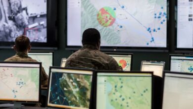 Two military personnel wearing camouflage uniform look at their computer screens displaying what appears to be maps. They're facing the background, which has three large screens displaying different maps. Similarly, four smaller screens make up the foreground.