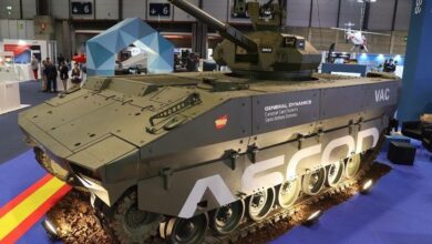 The Spanish Army's Vehículo de Apoyo de Cadenas (VAC) is seen on display in what appears to be a weapons expo, inside a large building with blue carpet. The tank is colored deep green.