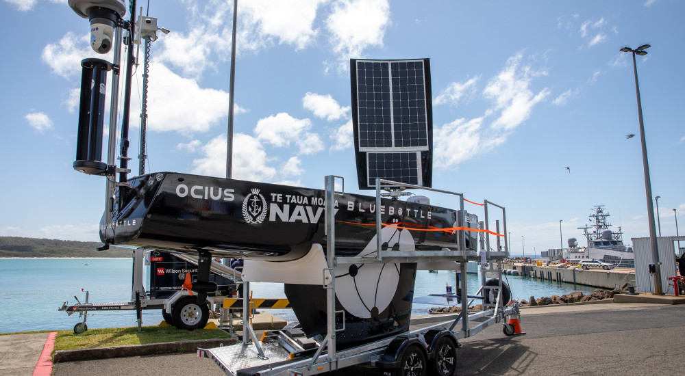 The Bluebottle is seen on a mobile wheeled platform, seemingly beign prepared for delivery. Its sail is propped up, with solar panels visibly attached to it. The black body of the vessel has "OCIUS", "Te Taua Moana Navy" (with their logo beside it), and "BLUEBOTTLE" painted on it in white. The background appears to be a port, with a grey ship on the far right side.