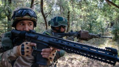 Two soldiers are seen patrolling in what appears to be a forest in daylight with their rifles at the ready. Both of them are pointing their weapons to the right. The one on the foreground, positioned on the left, is holding a Sig Sauer rifle. The other soldier is positioned behind his left, on the center of the image. They are both wearing camouflage.