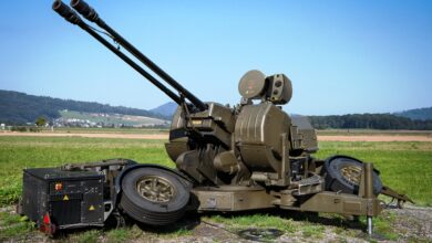 An Oerlikon GDF 103 anti-aircraft gun is seen set up in a grassy plains. Its two gun barrels are pointed upwards to the left at a 45-degree angle. The whole gun system is painted a deep green. The far background is a clear, blue sky.