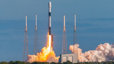 A Falcon 9 rocket launches from Cape Canaveral Space Force Station in Florida. Bright red-orange flames jut out from its back as it propels upward. Smoke is seen covering the lower right side of the image. The background is a calm blue sky.