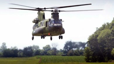 An medium-to-heavy lift CH-147F Chinook helicopter is seen hovering over a green landscape with trees sprawling in the background and the far right side of the image. The helicopter is colored a dark green and grey camouflage. Its front and back propellers are due to its fast rotations.The far background is a bright white sky.