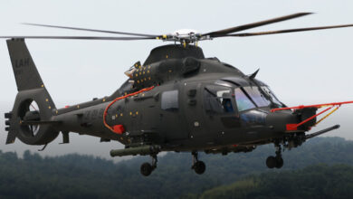 A dark green-colored light-armed helicopter is seen flying over a forest landscape.