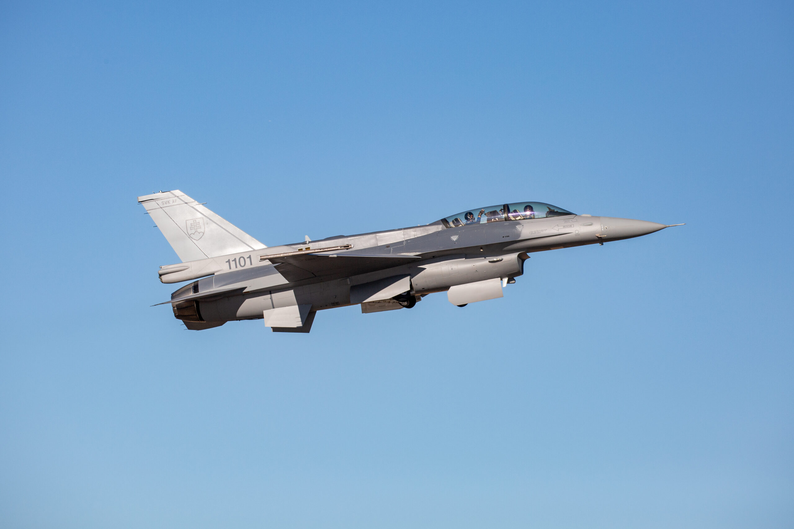 A gray Lockheed Martin F-16 jet plane is seen flying in clear blue skies. It has "1101" painted at the back, below its tail.