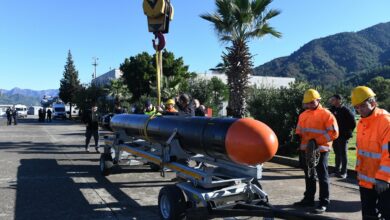 The AKYA torpedo is seen being placed on a wheeled cart. It is tied in the middle with a thick yellow rope, which is used by a crane's hook to place it gently on the cart. Around 17 people are seen scattered in the image, monitoring the weapon's movement. Four of them are wearing high-visibility jackets and hard hats. The area appears to be a military compound with lots of trees planted in the lot. The background is a green mountainous landscape. The sky is a clear and cool blue.