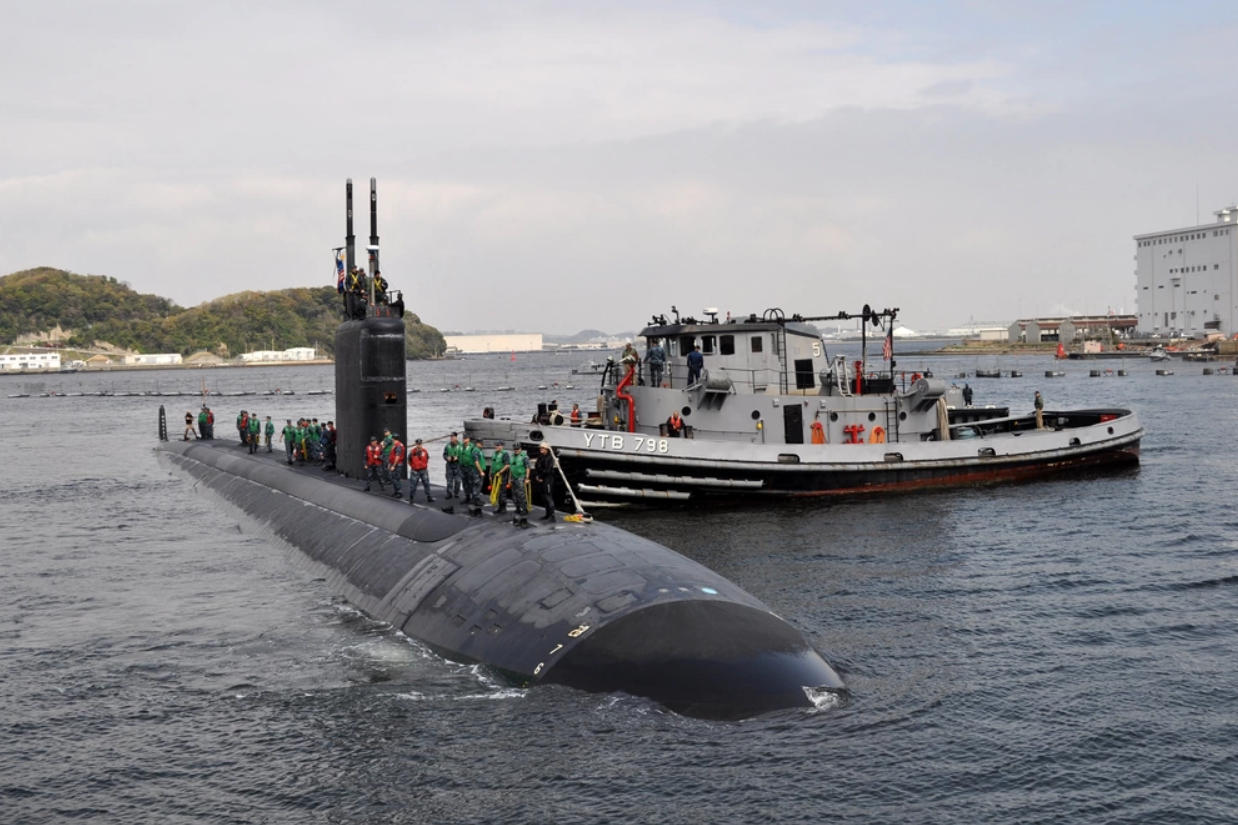 The USS Columbus submarine is seen partially submerged in the waters of what appears to be a pier. A boat with "YTB 798" painted on its side is seen right beside the black submarine. On top of its hull, around 25 people are seen standing, most of them wearing lifevests. In the background, a mountainous area is seen over the left of the horizon. On the right, two buildings that appear to be warehouses are also visible.