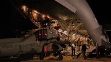 A gray AH-64E Version 6 Apache attack helicopter is seen being unloaded out from a US Air Force C-17 airlifter via a ramp. Ten individuals, seven of whom are wearing their camouflaged uniforms, are on the scene, occupied with the delivery of the aircraft.
