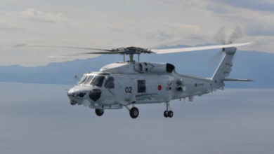 A Mitsubishi SH-60L is seen flying above the sea. The white-gray helicopter has "02" painted in black on its side. The background shows a mountainous shoreline, as well as the calm waters in its front. The sky is slightly gloomy and cloudy.