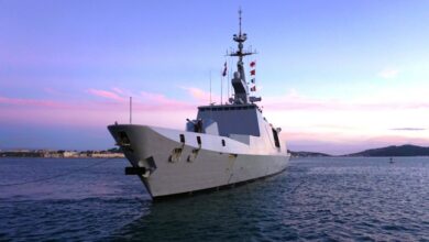 The French frigate Aconit (F713) is seen docked in what appears to be a pier. Its hull is painted gray. A rope is seen tied to its front. It has flags of varying colors decorated beside its mast. The water is a deep, dark blue. The background is a blue sky with purple clouds, reflecting the sun out of frame.
