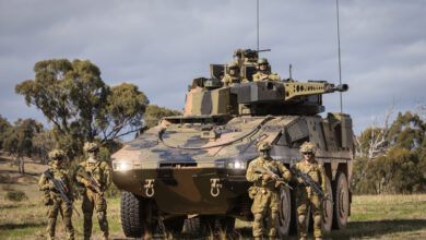 Rheinmetall Boxer Combat Reconnaissance Vehicle is seen with its headlights turned on in a grassy field. The armored 8x8 vehicle is painted in camouflage. Two pairs of camouflaged soldiers holding rifles flank its sides. On top of the vehicle, two more soldiers are seen. The sky is gray and filled with dark clouds, and some trees are seen scattered on the background.