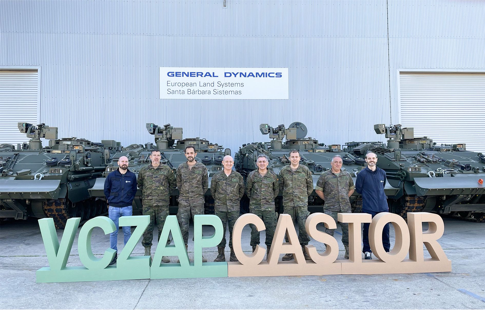 Six uniformed Spanish Army personnel stand in front of four ASCOD Castor armored combat vehicles. They are joined by two civilians, likely from General Dynamics. In front of them are letter standees that spell out "VCZAP CASTOR". The background is a warehouse building, with a sign that reads "GENERAL DYNAMICS European Land Systems Santa Barbara Sistemas".