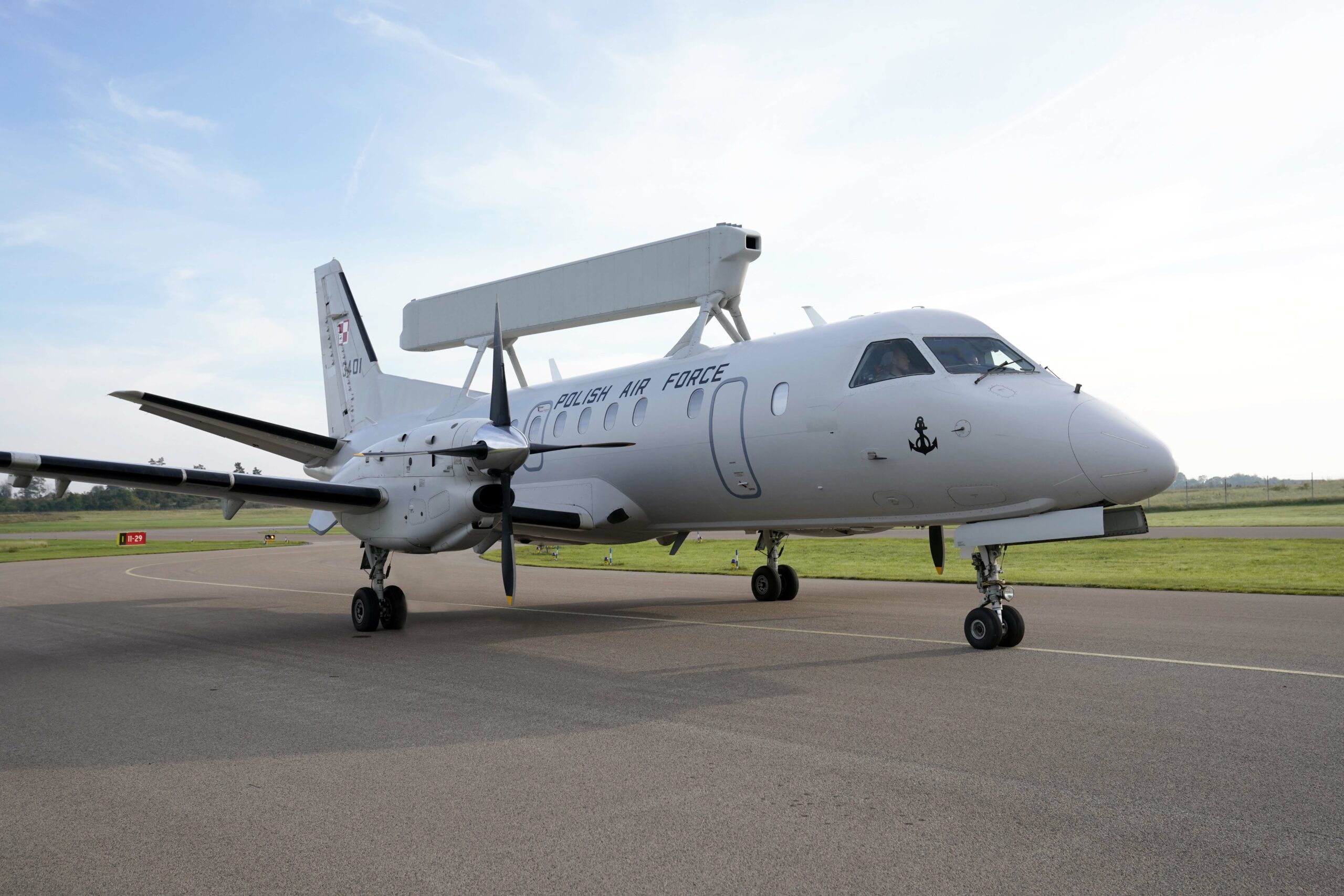Poland's Saab 340 Airborne Early Warning (AEW) aircraft is seen on a runway. The plane is white and has "POLISH AIR FORCE" painted on its right side. Its rectangular radar system is propped up above and perpendicular to its body. Its tail number, 3401, is seen on the right side of its tail.