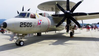 A Northrop Grumman E-2K Hawkeye plane appears to be on display at a wide-open area similar to a runway. It is cordoned off by a red-velvet stanchions seen in the back. The gray plane has "2505" painted on its left side in black, below the cockpit. It also has a red-white-blue logo with an eagle's head. Above it's body, its circular radar system is seen set parallel to its body.