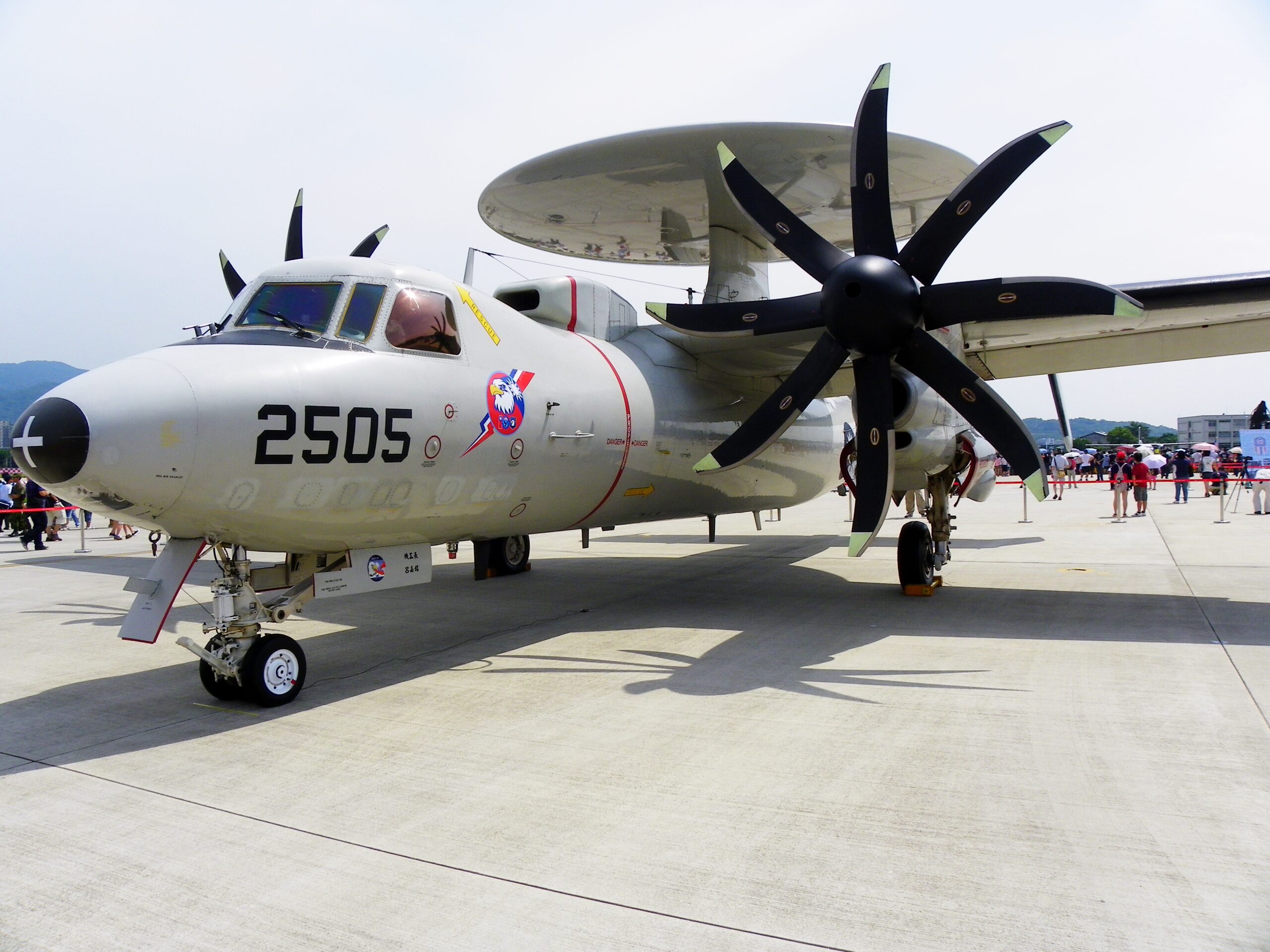 A Northrop Grumman E-2K Hawkeye plane appears to be on display at a wide-open area similar to a runway. It is cordoned off by a red-velvet stanchions seen in the back. The gray plane has "2505" painted on its left side in black, below the cockpit. It also has a red-white-blue logo with an eagle's head. Above it's body, its circular radar system is seen set parallel to its body.