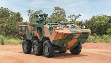 A Guarani 6x6 Armored Personnel Carrier is seen parked in an area with red sand and some trees and bushes in the background. The vehicle is painted in green and reddish-brown camouflage. A soldier wearing a helmet is seen peeking out the hatch. The far background is a blue sky with thin white clouds scattered evenly.