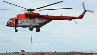Argentina's Mi-171E helicopter, H-95, is seen flying above a green plains. An orange cable with what appears to be a hook contraption on its end dangles from its body. The helicopter is painted orange on its upper half and outside the cockpit, and gray on its lower half. Its body has many writings and decals on it, including "H-95" and "Fuerza Aérea Argentina" painted in black and all caps. Below, on the ground, an individual wearing an orange high-vis jacket and black pants appear to be looking up the aircraft. Close to him, on the foreground, are dozens of orange container barrels.