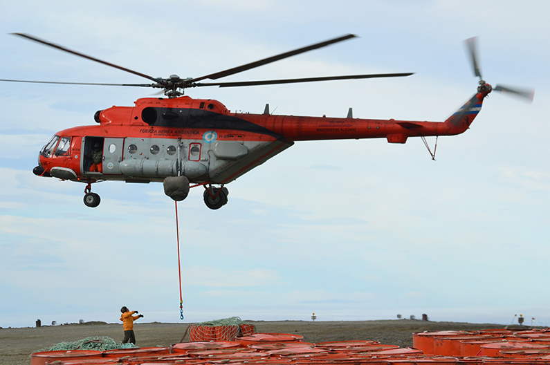Argentina's Mi-171E helicopter, H-95, is seen flying above a green plains. An orange cable with what appears to be a hook contraption on its end dangles from its body. The helicopter is painted orange on its upper half and outside the cockpit, and gray on its lower half. Its body has many writings and decals on it, including "H-95" and "Fuerza Aérea Argentina" painted in black and all caps. Below, on the ground, an individual wearing an orange high-vis jacket and black pants appear to be looking up the aircraft. Close to him, on the foreground, are dozens of orange container barrels.