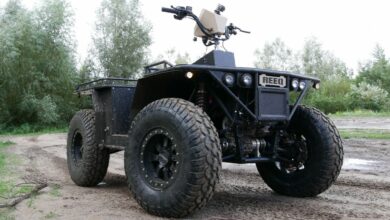 A REEQ Rex Quad is seen parked in a muddy field with trees and shrubbery in the background. The vehicle is a 4-wheel off-road vehicle with large wheels and a minimalistic chassis. Most of its parts are painted black, except for the logo in front and the casing of the screen in the middle of the handlebars, which are both dirty brown.