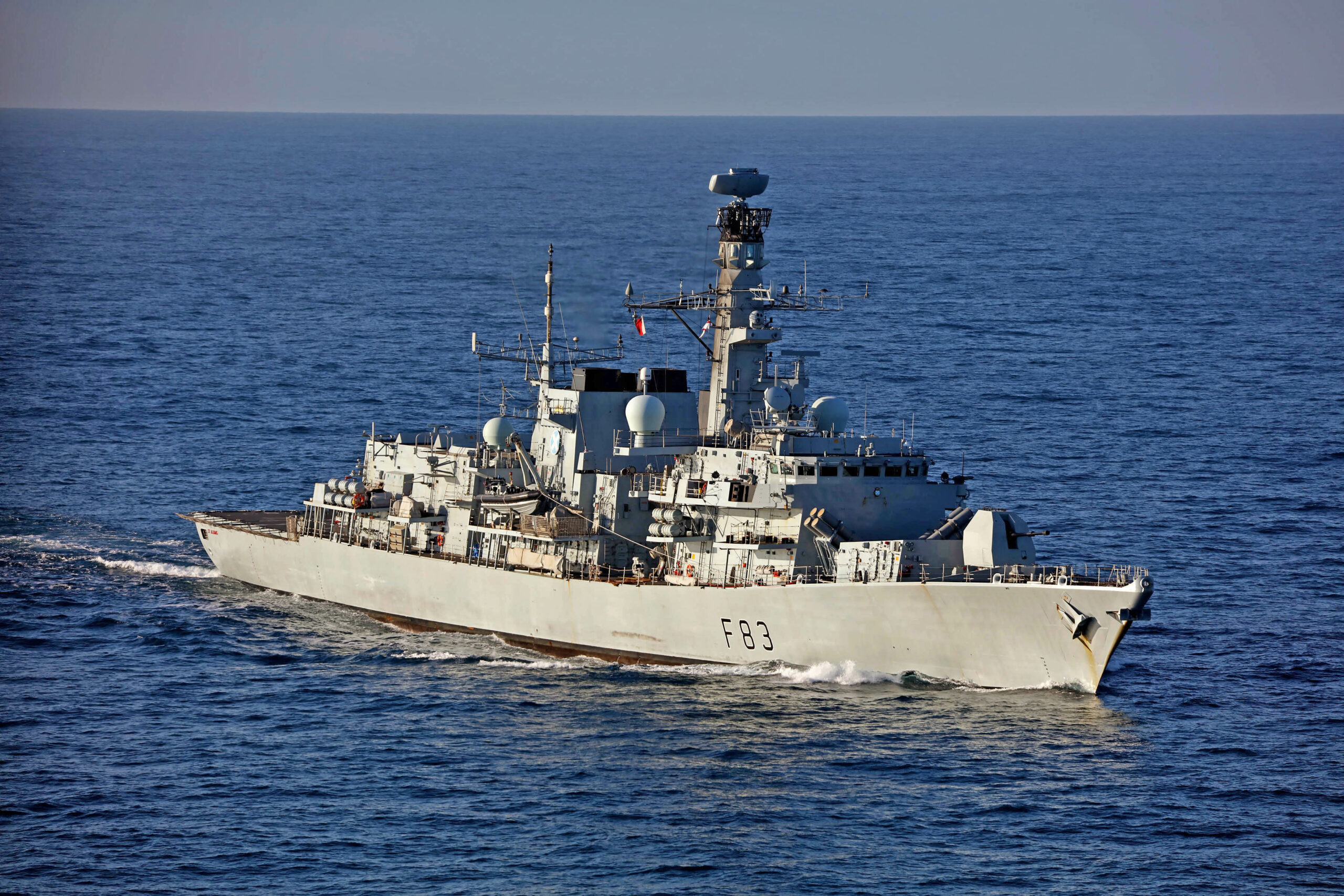 The UK Royal Navy's HMS St Albans frigate is seen sailing in calm, deep blue waters. The ship is painted gray, with "F83" painted on the right side of its hull in black. The far background is a gray sky.