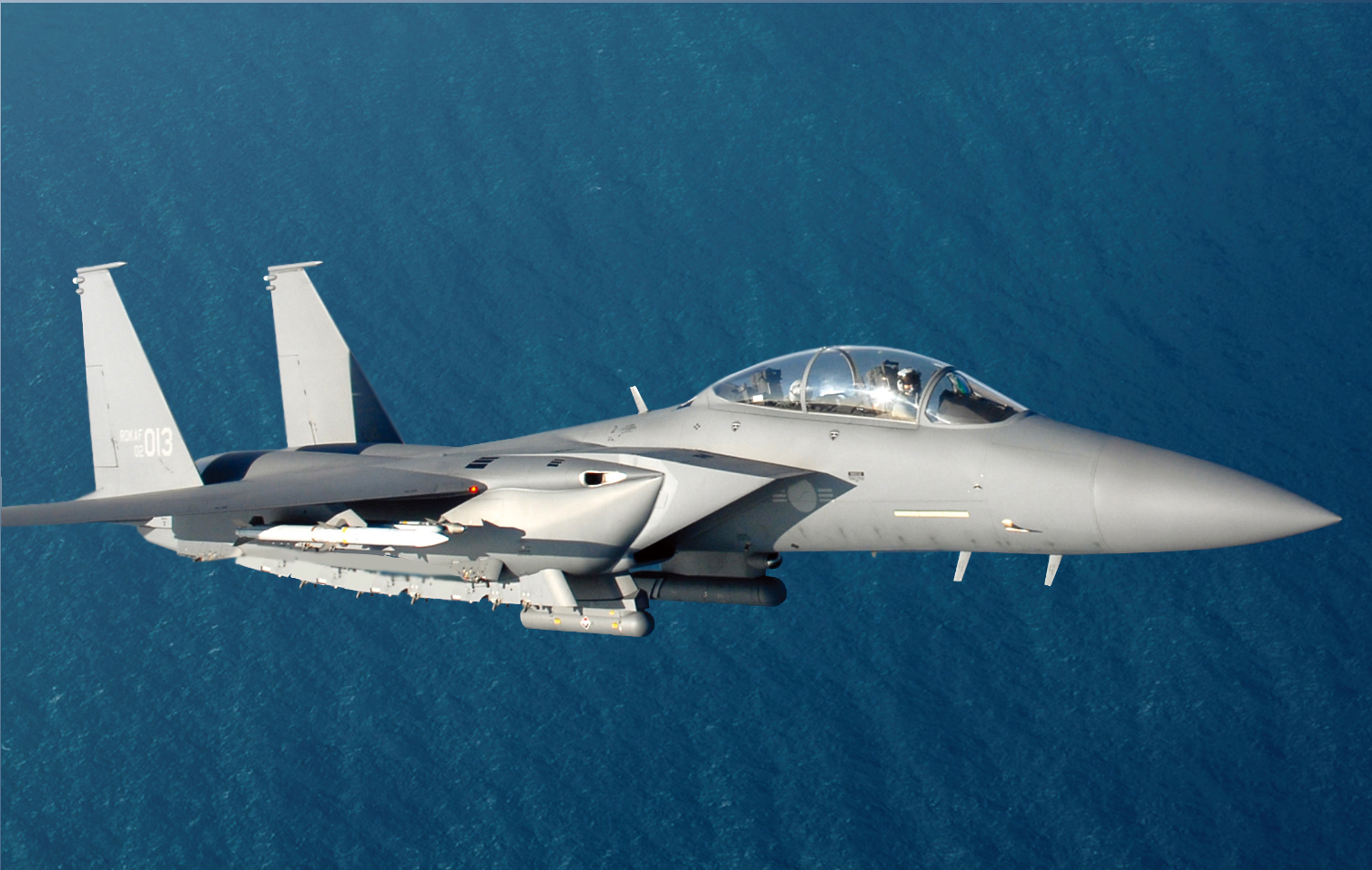 A Republic of Korea Air Force F-15K fighter plane is seen flying above a body of water. The fighter plane is painted gray and has a sleek body to make it more aerodynamic. The number "013" is painted on its tail in white.