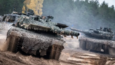 Three armored tanks are seen moving in a sandy area with trees in the back. The tanks are covered with ghillie suit fabric to help with camouflaging.