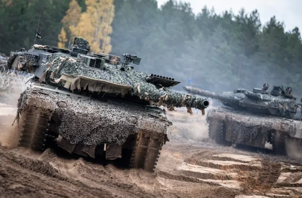 Three armored tanks are seen moving in a sandy area with trees in the back. The tanks are covered with ghillie suit fabric to help with camouflaging.